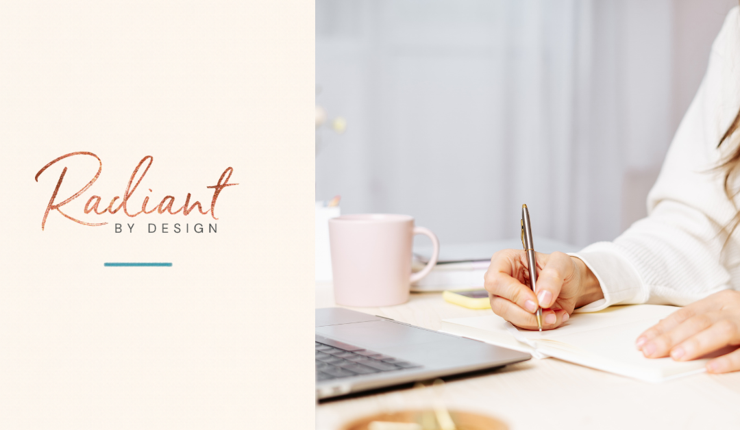 A woman in a white sweater writes in a notebook at a desk beside a pink mug and an open laptop.