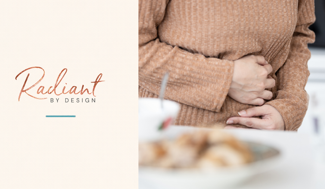 A woman in a tan ribbed sweater holds her stomach with both hands beside a blurred plate of food.