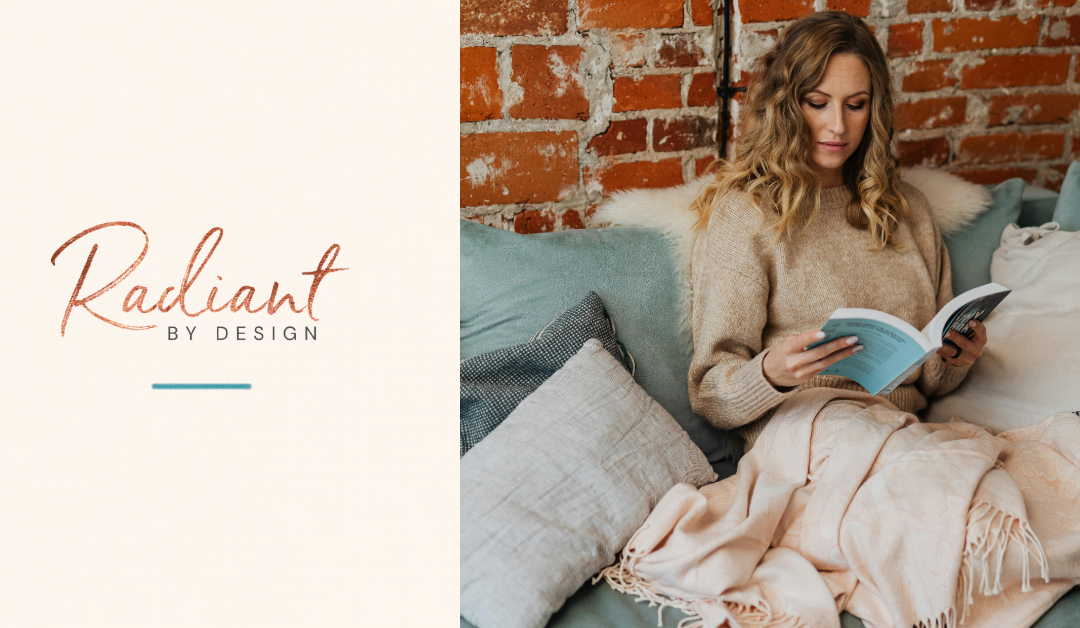 A woman with wavy blonde hair reads a paperback on a sage-green sofa in front of a brick wall, with a blush blanket over her lap.
