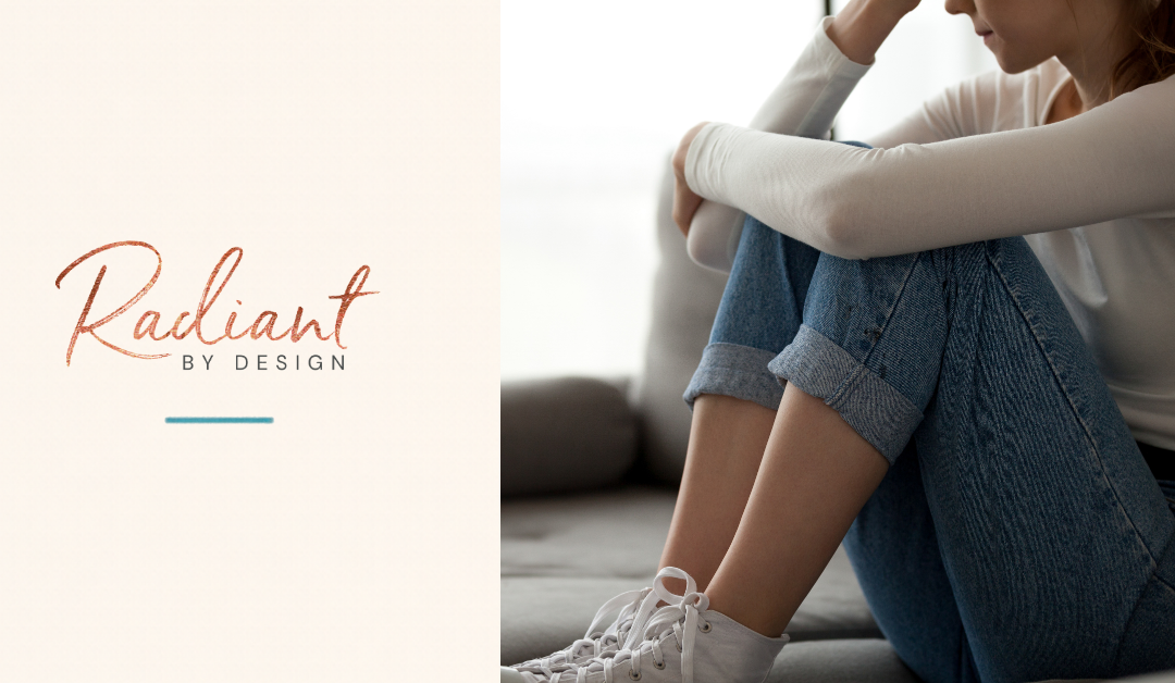 A woman in a cream top and rolled jeans sits on a gray couch with her knees pulled up and her head resting on her arm.