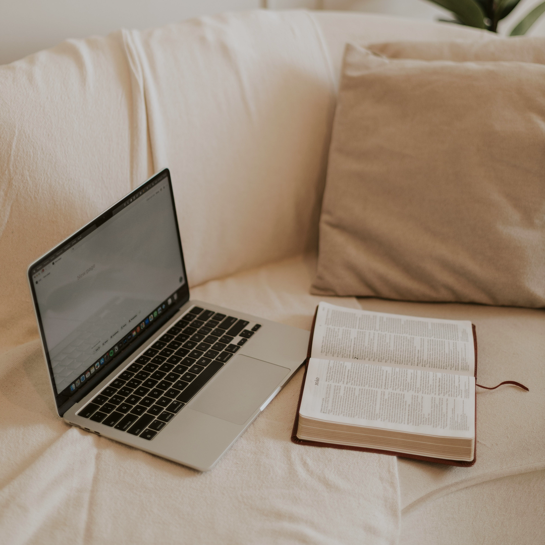 Laptop and an open Bible resting on a cream sofa beside a linen cushion.